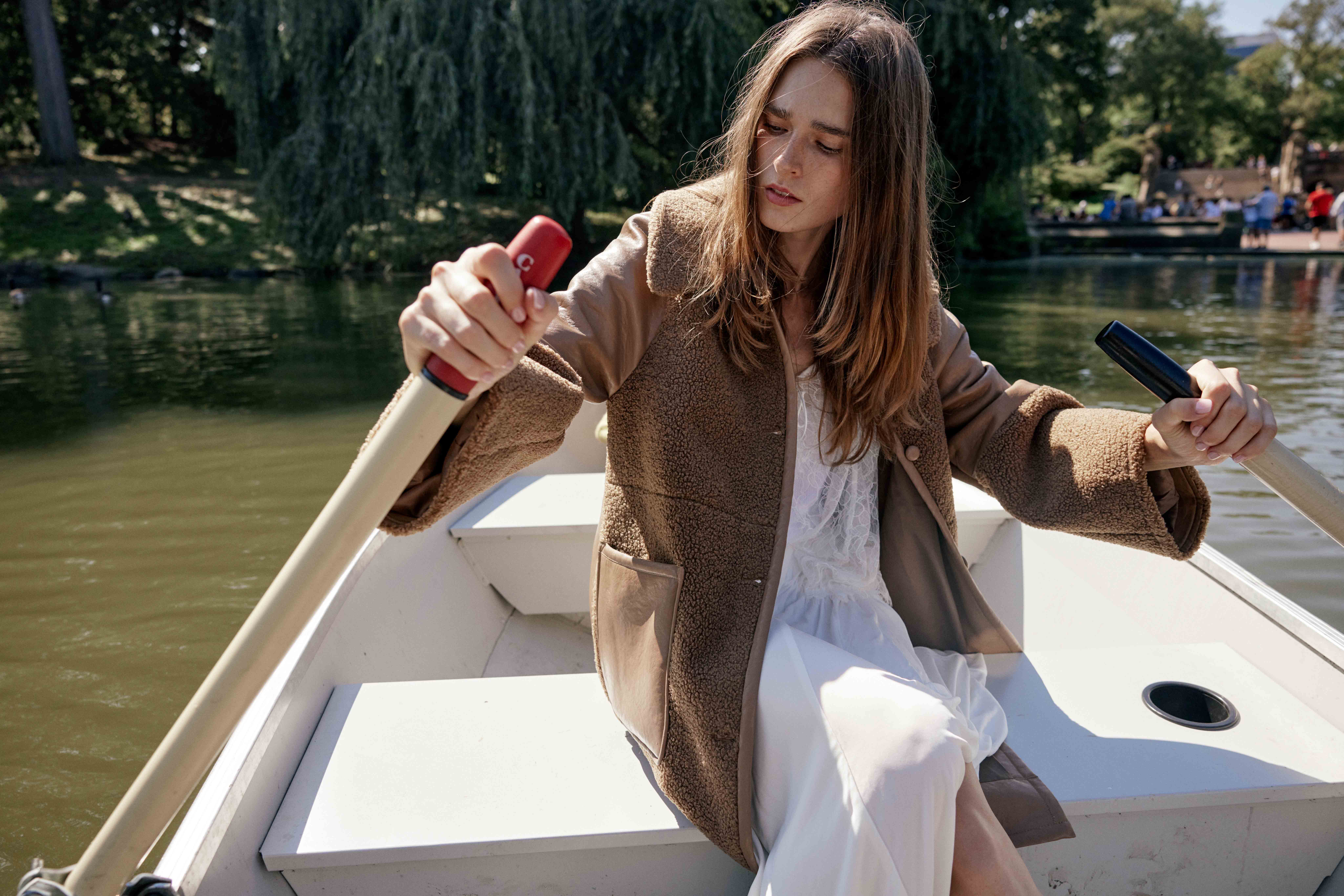 A fair-skinned woman with long brown hair rows a boat on a lake, wearing a teddy bear coat and white dress, surrounded by trees.