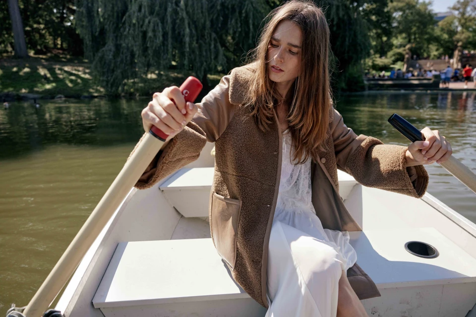 A fair-skinned woman with long brown hair rows a boat on a lake, wearing a teddy bear coat and white dress, surrounded by trees.