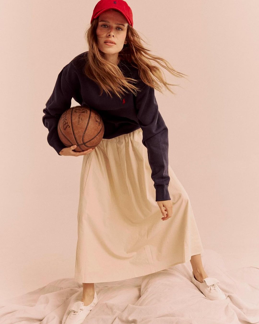 Woman in baseball cap poses with a basketball in a studio setting, wearing a navy sweater and cream-colored skirt and sneakers.