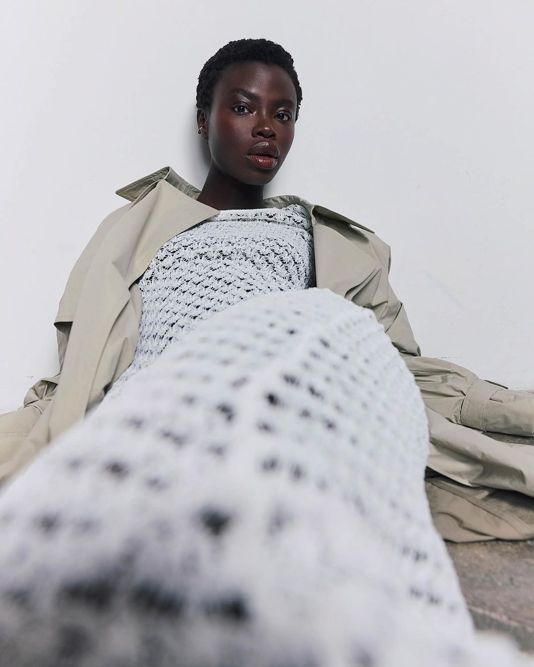 Fashion portrait of a dark-skinned woman wearing a white crochet top and tan trench coat, sitting against a white backdrop.