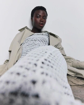 Fashion portrait of a dark-skinned woman wearing a white crochet top and tan trench coat, sitting against a white backdrop.