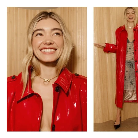 Blonde woman in a red coat with a choker necklace smiles against a light wooden wall with vertical stripes. Fashion editorial shot.