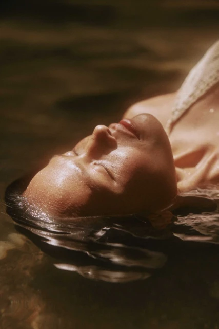 Woman with closed eyes and wet dark hair floats in a body of water, possibly a lake or pool, in warm sunlight in a medium shot.