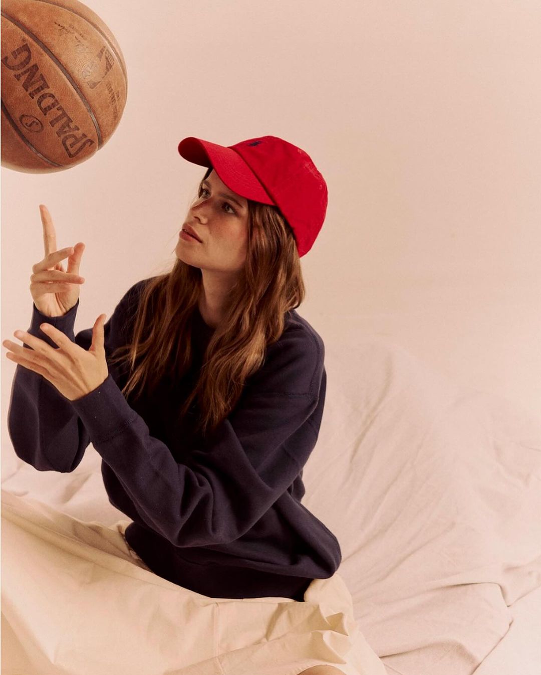 Young adult woman in red baseball cap seated, casually tossing a basketball in an indoor setting with soft, diffused lighting.