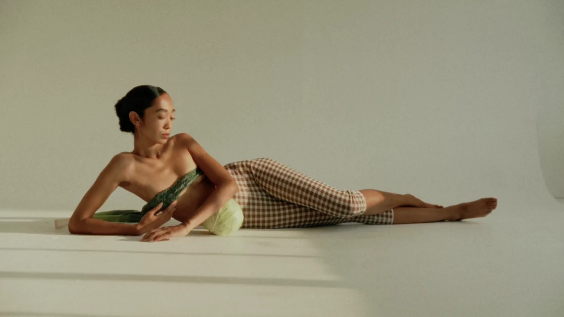 Asian woman reclines in studio setting with vegetables including cucumber and cabbage in an editorial-style, warm-toned composition.