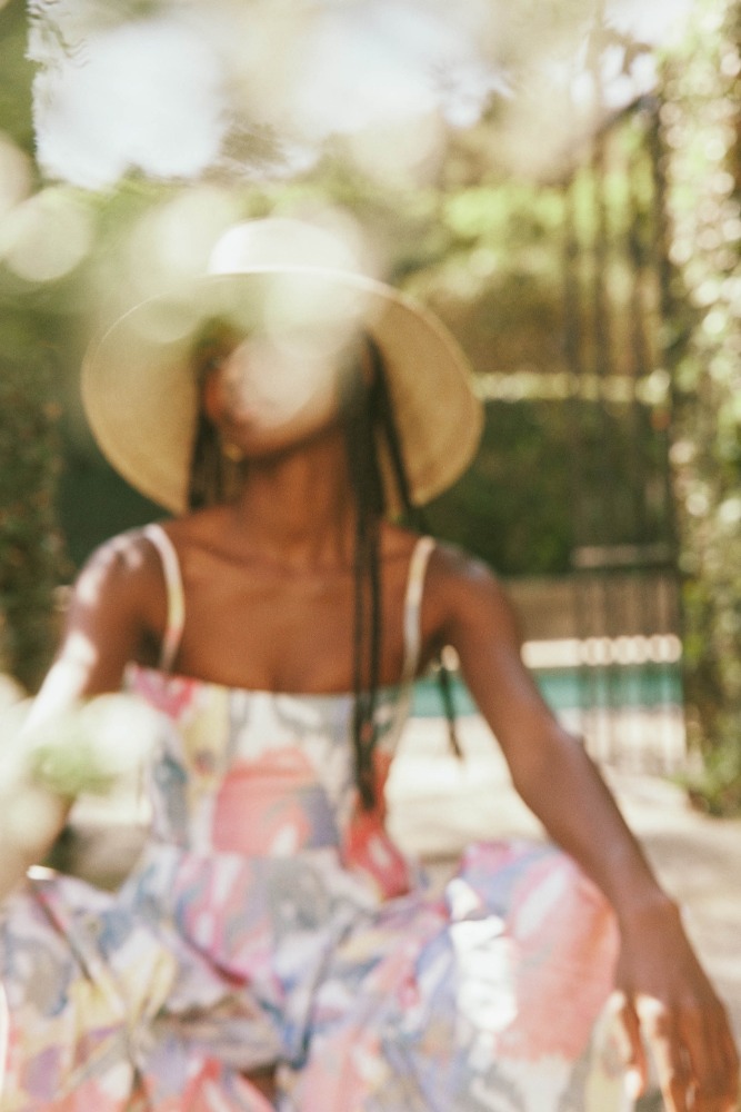 Woman wearing a wide-brimmed hat and patterned dress outdoors, captured through a soft-focus foreground foliage layer.