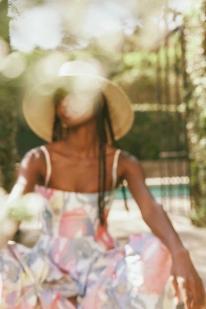 Woman wearing a wide-brimmed hat and patterned dress outdoors, captured through a soft-focus foreground foliage layer.