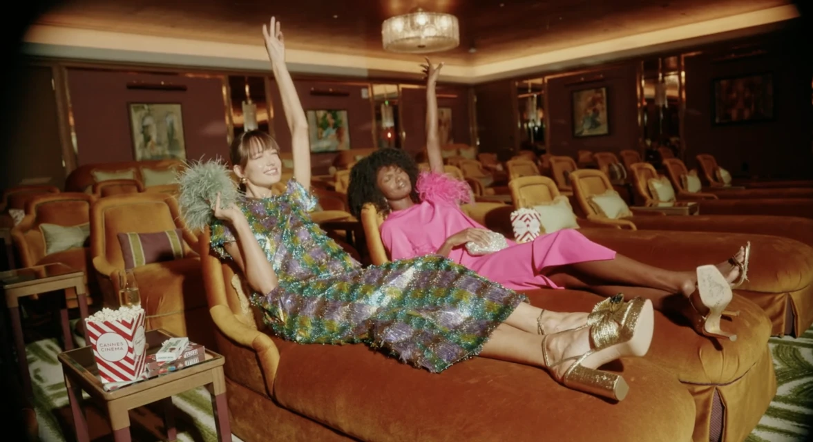Two women in colorful dresses lounge in a vintage-style movie theater, arms raised, with popcorn and accessories, bathed in warm light.
