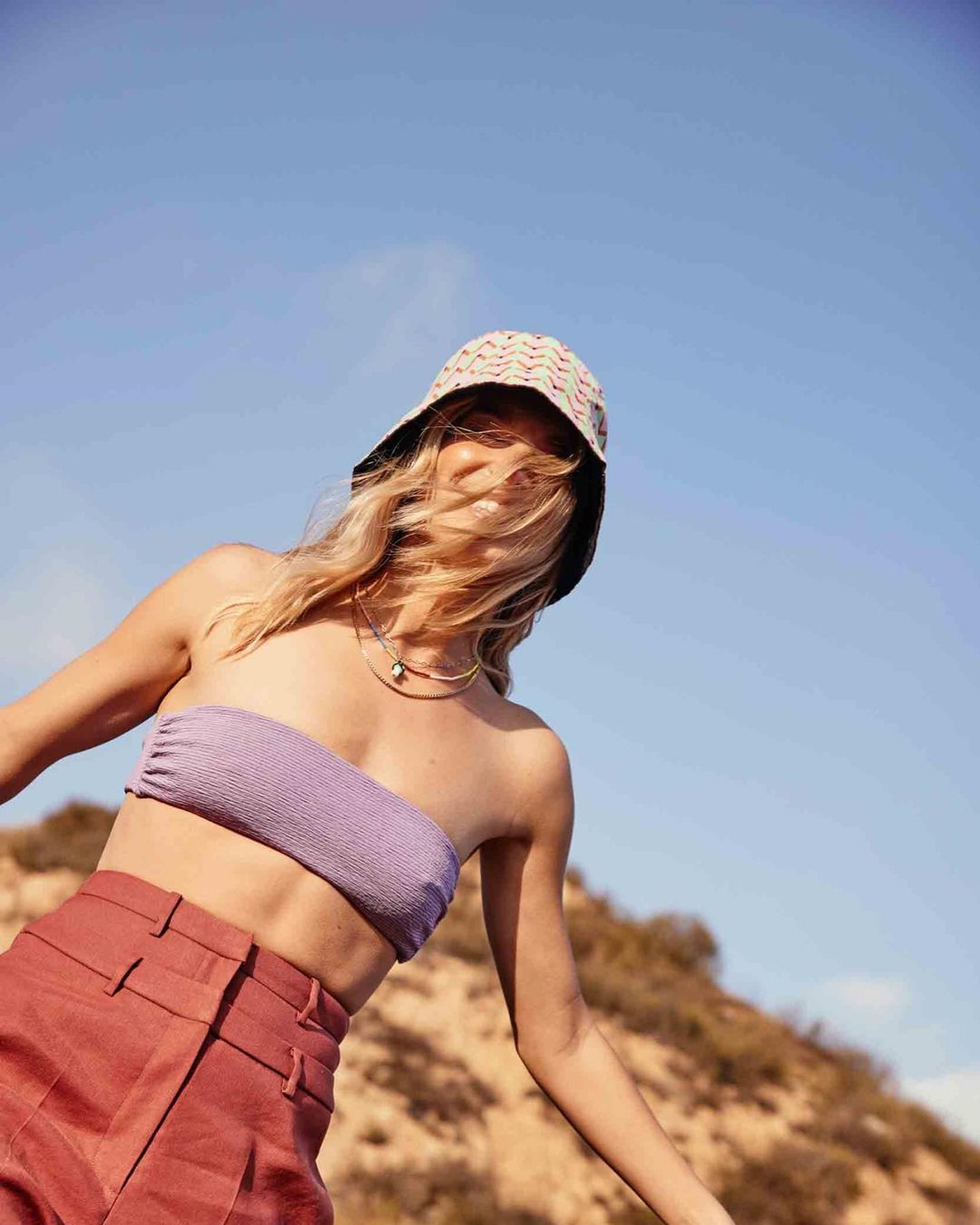 Woman in a bucket hat, purple tube top, and red pants poses in a sunny outdoor environment. Fashion and lifestyle photography.