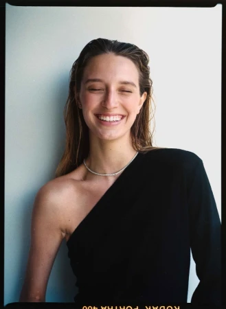 Portrait of a smiling woman with wet hair wearing a black one-shoulder top and a delicate silver necklace against a plain wall.