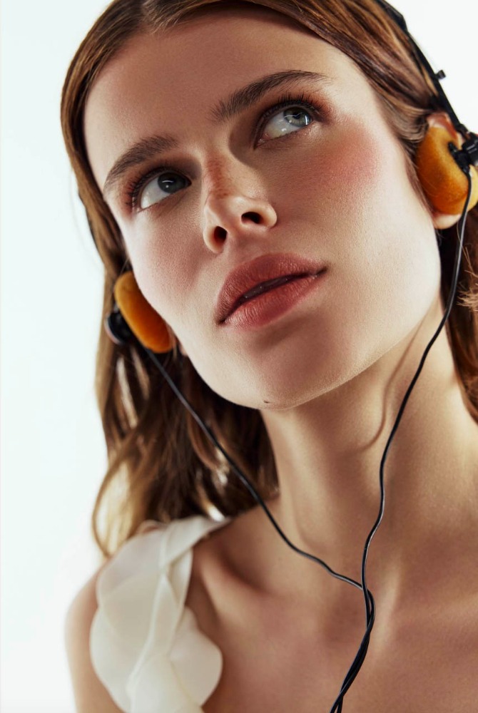 Close-up portrait of a woman with brown hair, wearing headphones, looking up with a soft, dreamy gaze against a white background.