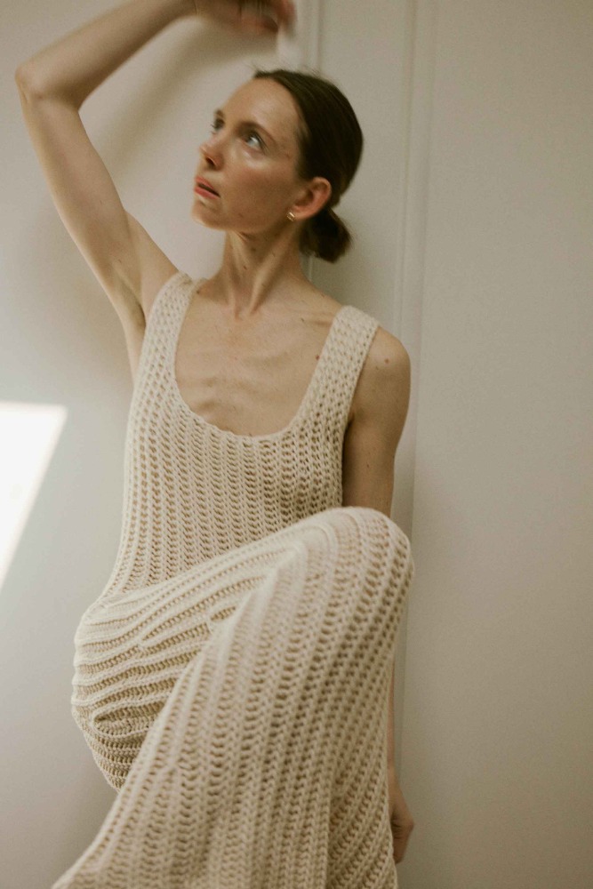 Woman in crocheted dress sitting against a wall, looking up with natural makeup in an indoor setting with diffused lighting.