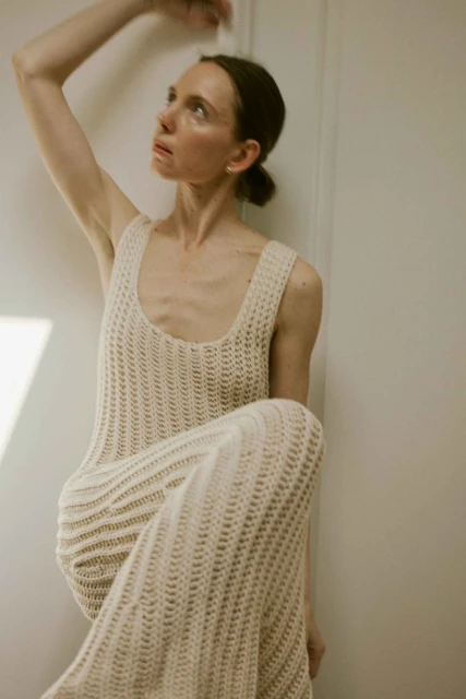 Woman in crocheted dress sitting against a wall, looking up with natural makeup in an indoor setting with diffused lighting.