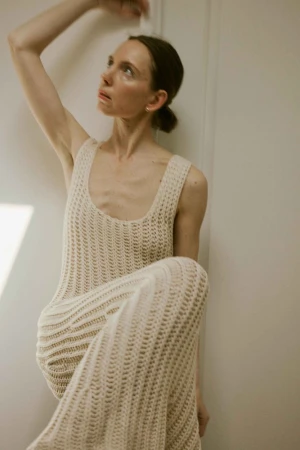 Woman in crocheted dress sitting against a wall, looking up with natural makeup in an indoor setting with diffused lighting.
