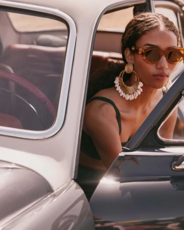 Woman in sunglasses wearing statement earrings sits in a classic car. Fashion and automotive elements are captured in natural light.