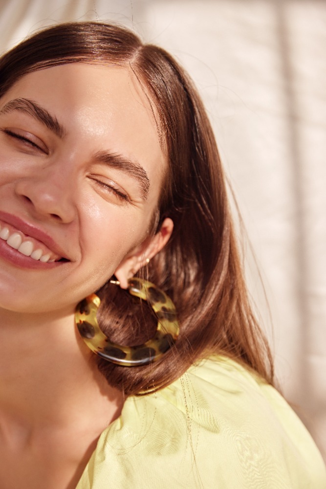 Close-up portrait of a smiling woman with brown hair wearing tortoiseshell hoop earrings and a yellow top in natural sunlight