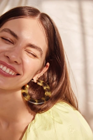 Close-up portrait of a smiling woman with brown hair wearing tortoiseshell hoop earrings and a yellow top in natural sunlight