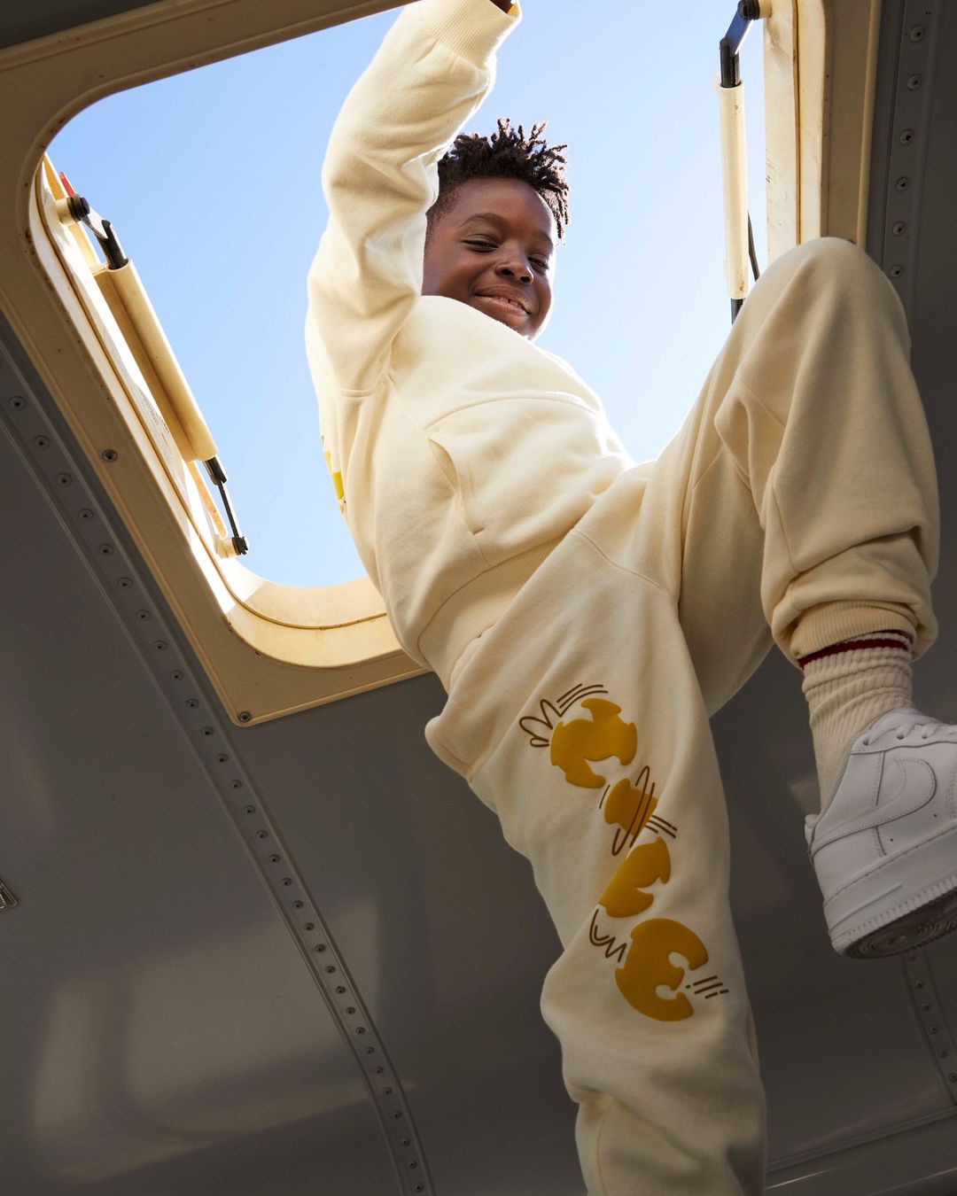 Low-angle view of a smiling Black child climbing through an opening, wearing a cream-colored sweatsuit and white sneakers against a blue sky.