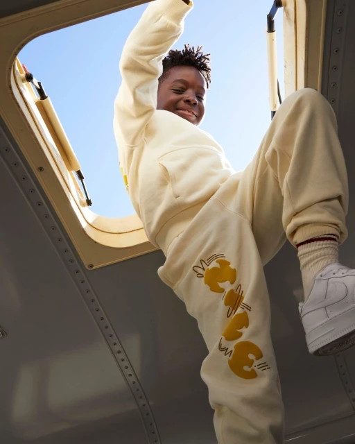 Low-angle view of a smiling Black child climbing through an opening, wearing a cream-colored sweatsuit and white sneakers against a blue sky.