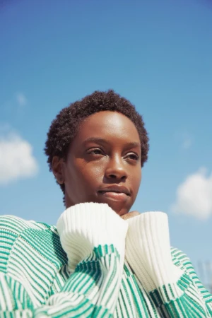 Portrait of a Black woman in her 20s wearing a green and white striped sweater against a bright blue sky.