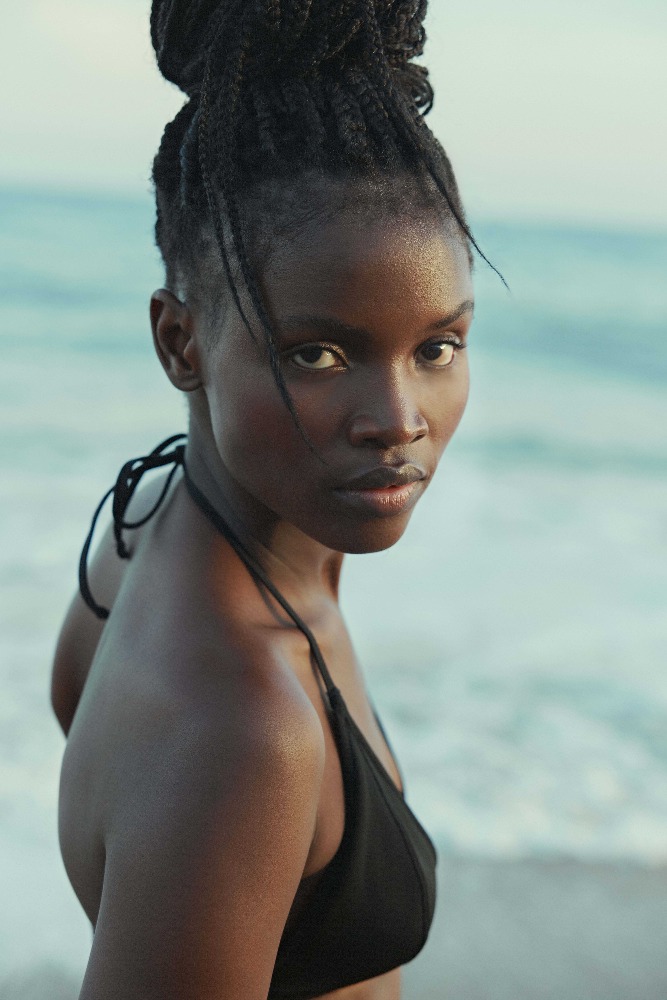 Close-up portrait of an African-American woman with braided hair wearing a black bikini top against a beach and ocean backdrop.
