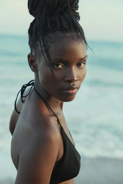 Close-up portrait of an African-American woman with braided hair wearing a black bikini top against a beach and ocean backdrop.