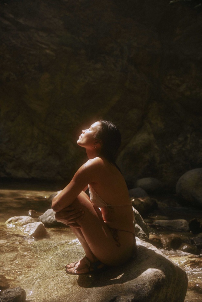 Woman crouched on a wet rock beside a stream, basking in bright sunlight in a natural rocky environment.