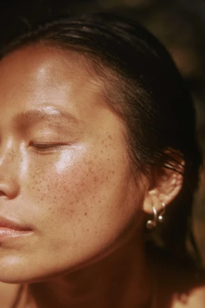 Close-up portrait of a woman with wet dark hair and freckles across her cheeks wearing small silver hoop earrings by Kate Moore.