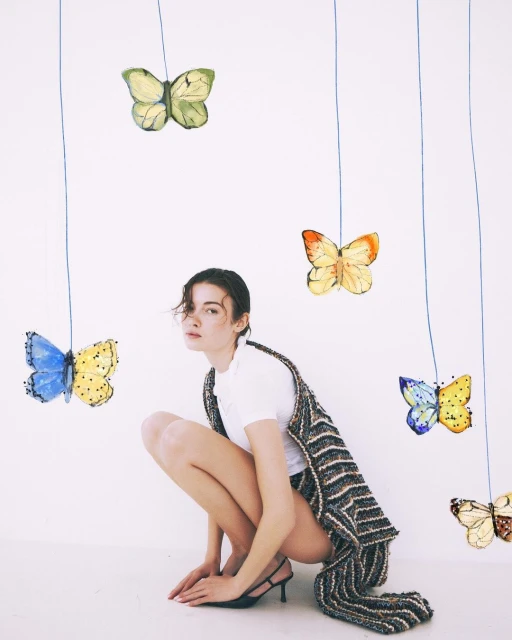 Young brunette woman squats indoors wearing a striped vest, butterflies dangle from strings against a white background in a fashion setting.