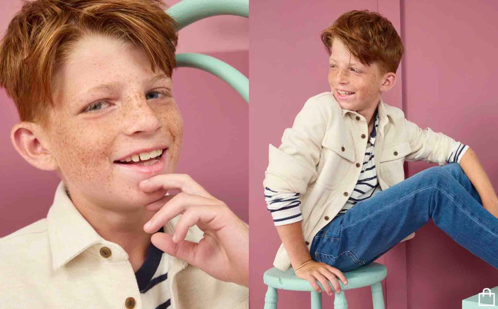 Boy with red hair and freckles poses in front of a pink wall while wearing jeans and a white jacket, sitting on a green stool.