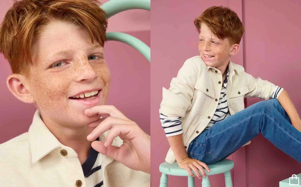 Boy with red hair and freckles poses in front of a pink wall while wearing jeans and a white jacket, sitting on a green stool.