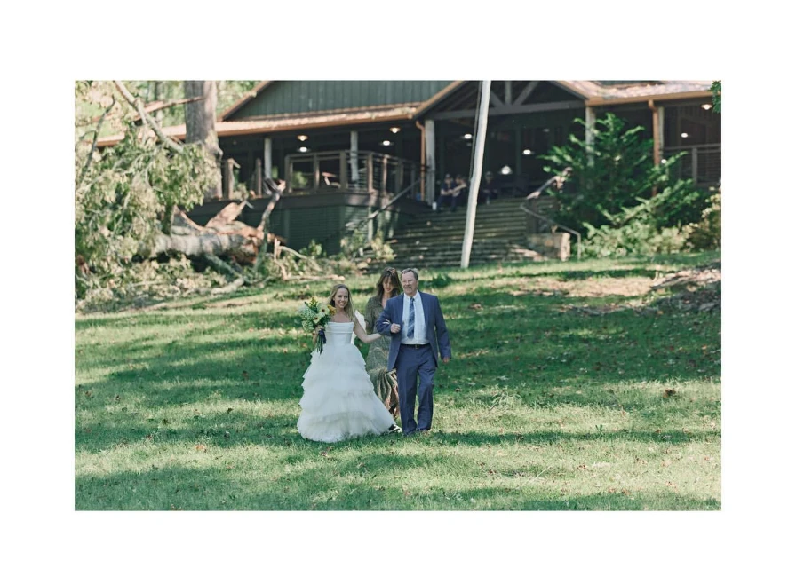 Bride and father walk across a grassy lawn with a bouquet during an outdoor wedding, with a building and trees in the background.