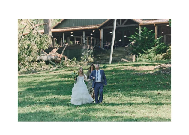 Bride and father walk across a grassy lawn with a bouquet during an outdoor wedding, with a building and trees in the background.