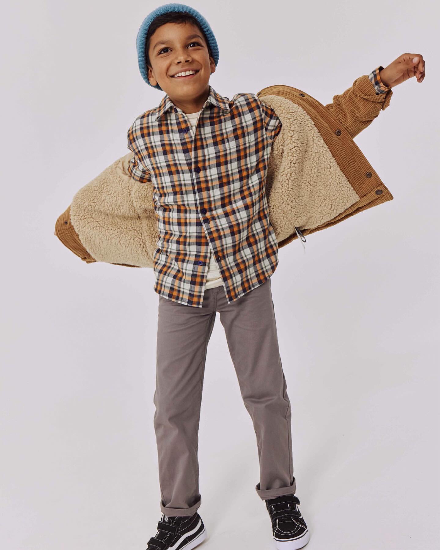 Smiling boy in a blue beanie, brown plaid shirt, brown pants, and black shoes with an open brown sherpa jacket, studio shot.