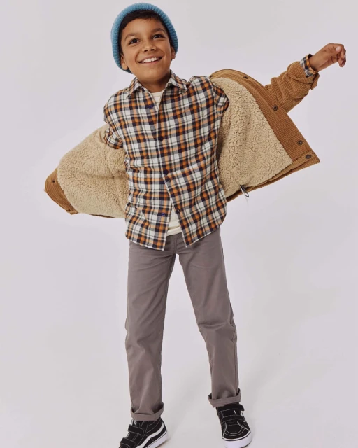 Smiling boy in a blue beanie, brown plaid shirt, brown pants, and black shoes with an open brown sherpa jacket, studio shot.