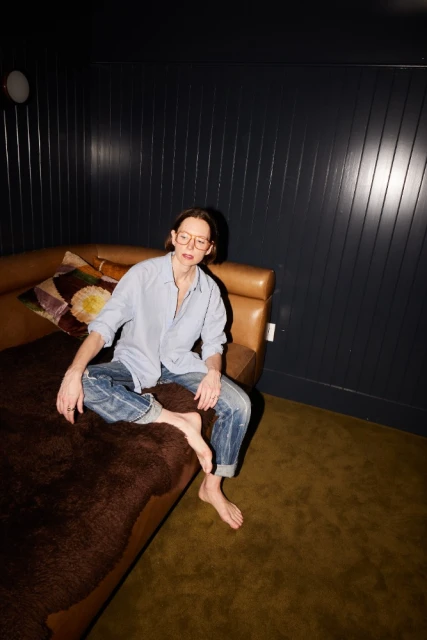 A young adult sits barefoot on a brown couch in a room with dark blue walls, wearing a light blue shirt and cuffed blue jeans.