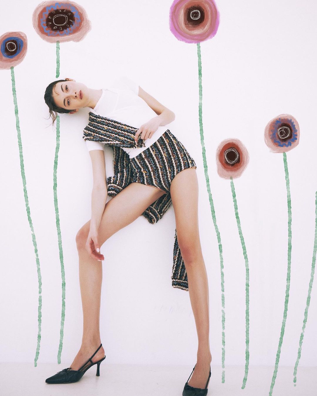 A slender woman with dark hair poses in striped shorts and heels against a backdrop of simple, painted flower stems and blooms.