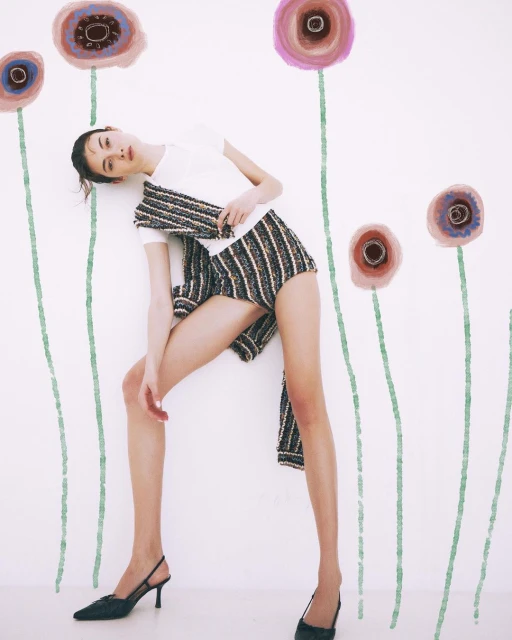 A slender woman with dark hair poses in striped shorts and heels against a backdrop of simple, painted flower stems and blooms.