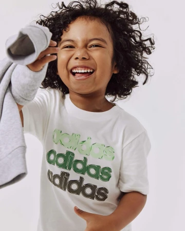 Smiling child with curly hair wearing a white t-shirt with green logo lettering against a white background