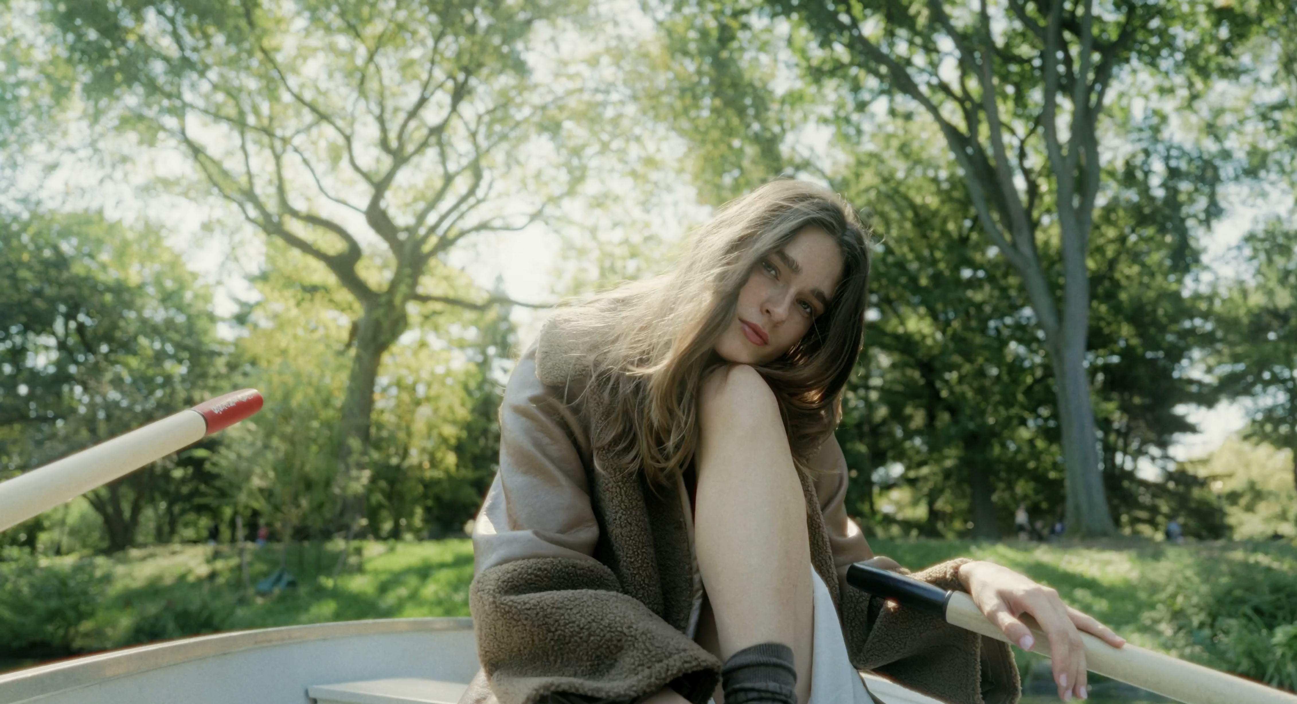 Young woman with long brown hair poses in a rowboat on a lake surrounded by green trees on a sunny day, wearing a brown coat.