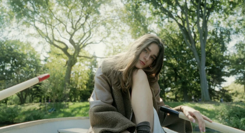 Young woman with long brown hair poses in a rowboat on a lake surrounded by green trees on a sunny day, wearing a brown coat.