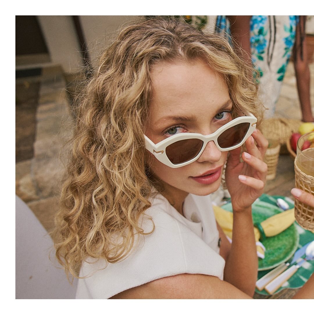 Close-up of a blonde woman with white-framed sunglasses, seated at an outdoor table with a casual, sunny aesthetic.