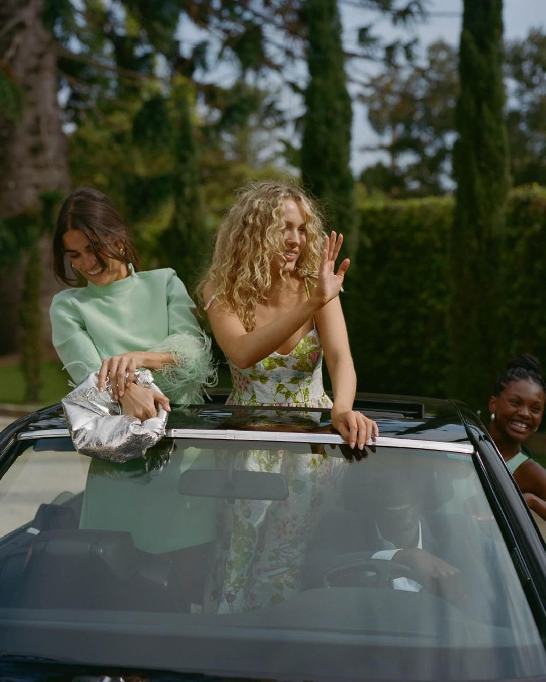 Three women are standing in a convertible car with the top down, smiling, on a sunny day with trees in the background.