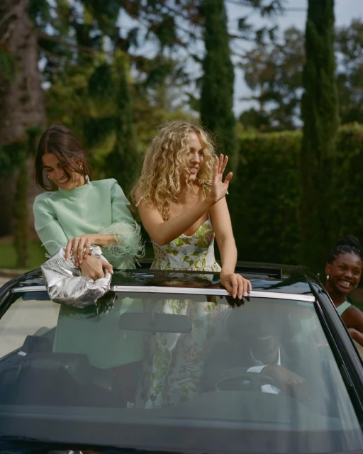 Three women are standing in a convertible car with the top down, smiling, on a sunny day with trees in the background.