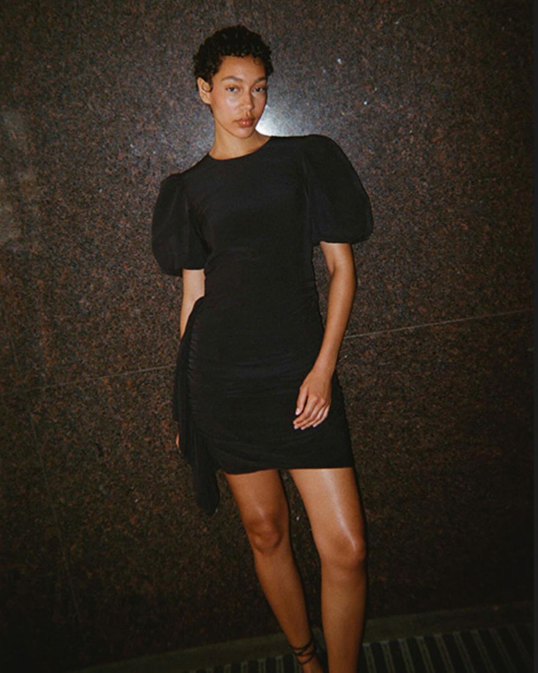 A stylish woman poses in a short black dress with puff sleeves in front of a textured wall, showcasing a blend of elegance and fashion.
