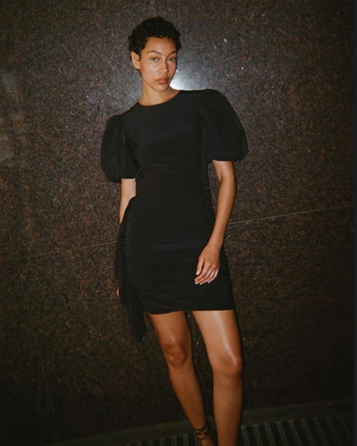 A stylish woman poses in a short black dress with puff sleeves in front of a textured wall, showcasing a blend of elegance and fashion.