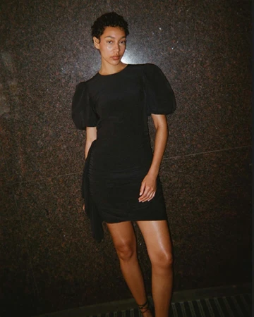 A stylish woman poses in a short black dress with puff sleeves in front of a textured wall, showcasing a blend of elegance and fashion.