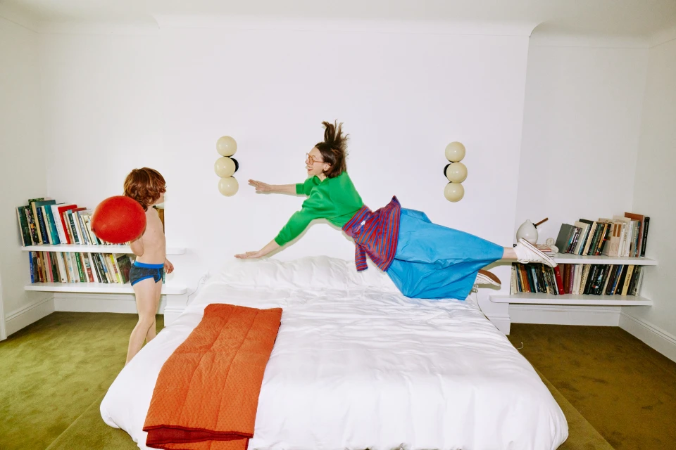 Woman in mid-air leaping above a bed, child with red ball watches in bedroom with white walls, shelves of books, and green carpet.