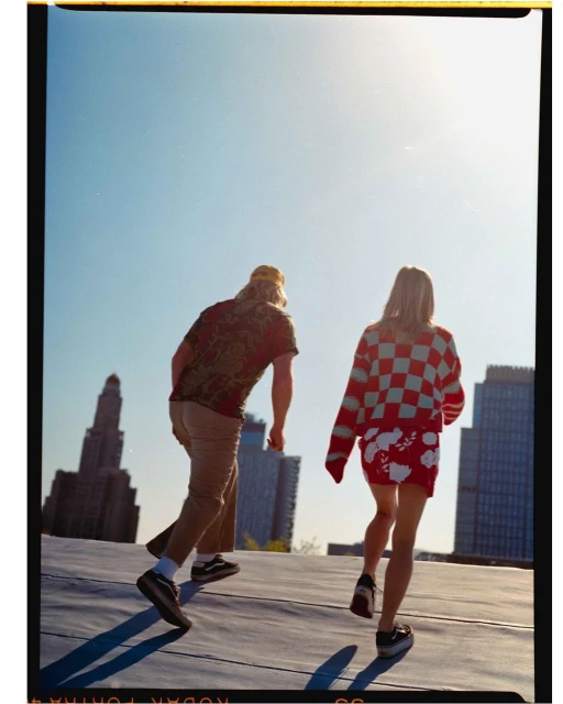 Two people walk on a rooftop on a sunny day with skyscrapers in the background. A man and woman are from behind, wearing casual clothing.