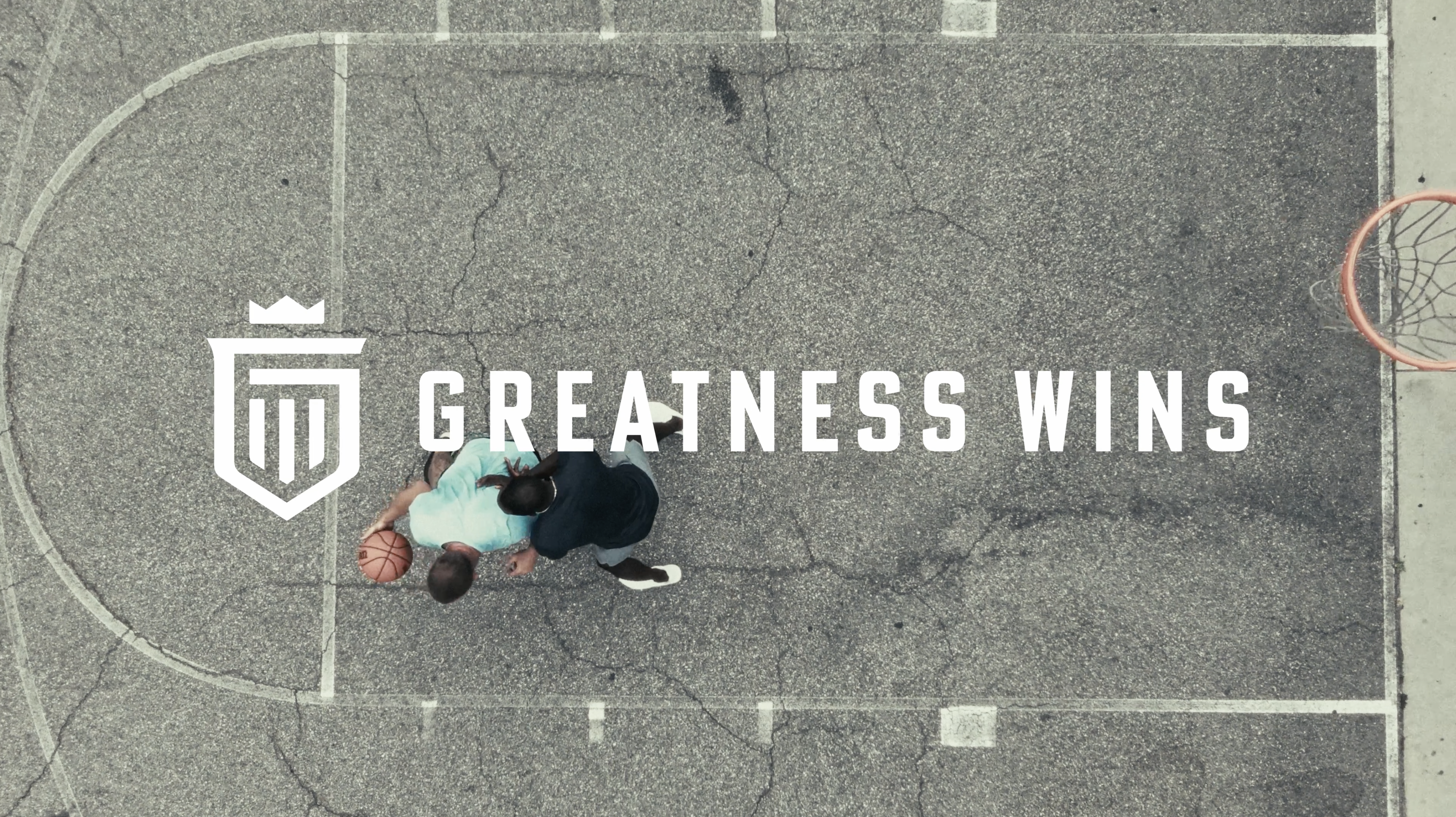 Overhead view of two adults playing basketball on an outdoor court with the text "Greatness Wins" and a white logo.